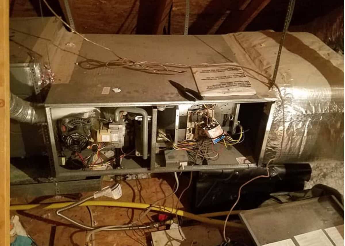 An open horizontal gas furnace installed in an unfinished attic, with the access panel removed to reveal internal wiring, control board, and blower motor components.